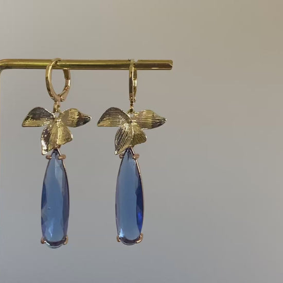 Filmpje van 14K vergulde en handgemaakte oorbel met vergulde oorringetjes, blaadjes en een langwerpige blauwe rechthoekige glasbedel. 