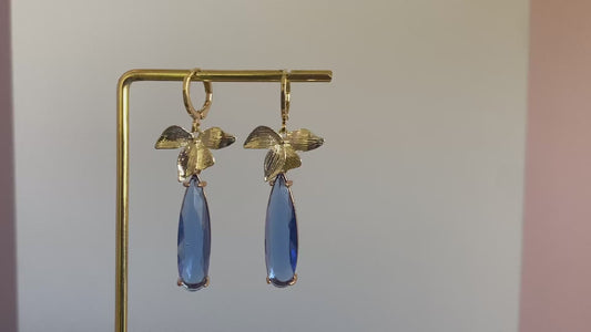 Filmpje van 14K vergulde en handgemaakte oorbel met vergulde oorringetjes, blaadjes en een langwerpige blauwe rechthoekige glasbedel. 